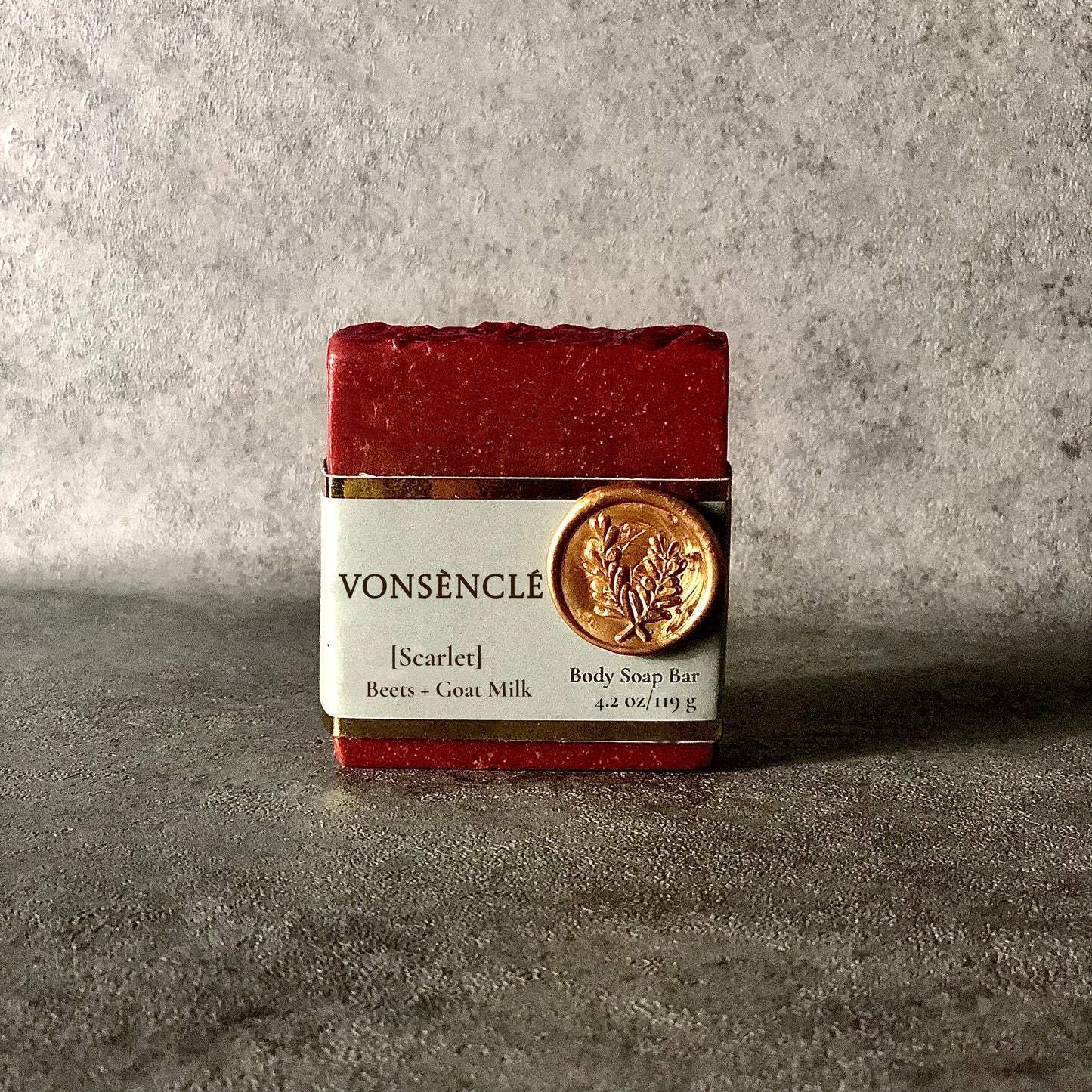 A Burgundy-red soap bar, wrapped with a grey-colored gold foil label.
