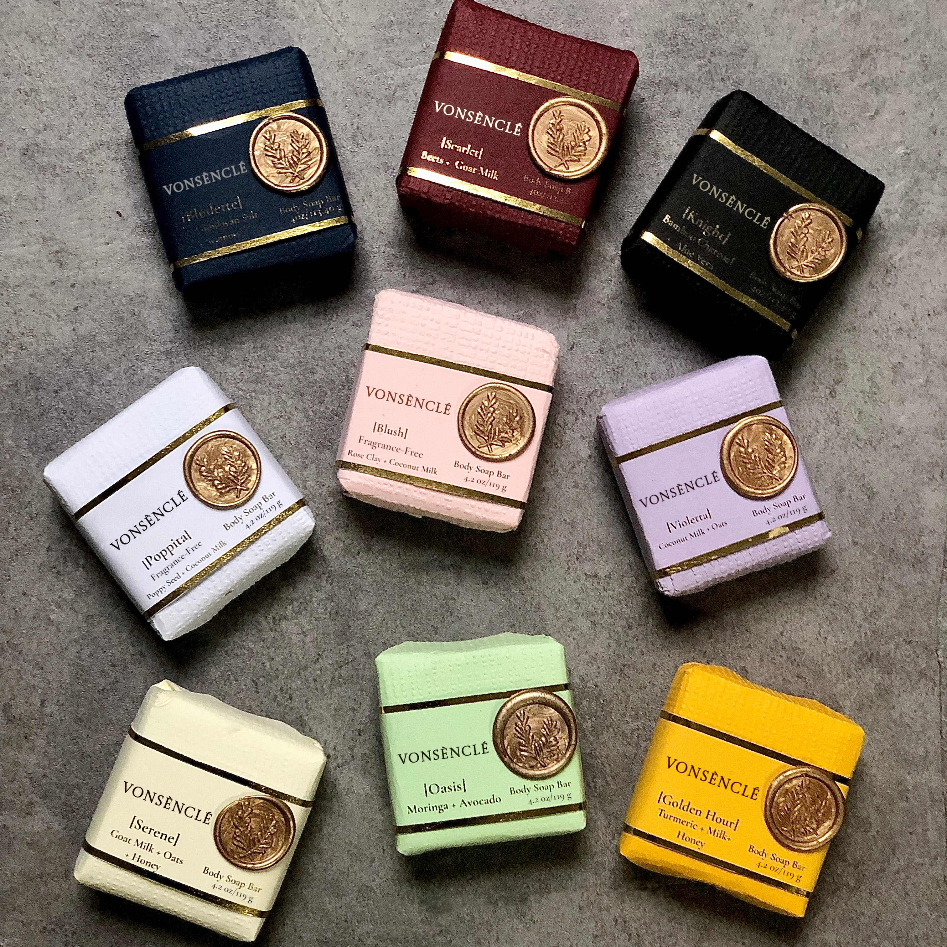 Eight packaged Vonsencle soap bars arranged in a circle on a gray textured surface. The packaged soap come in assorted colors including navy, burgundy, black, white, pink, lavender, light green, cream, and yellow, each with a gold seal and the Vonsencle brand name visible.
